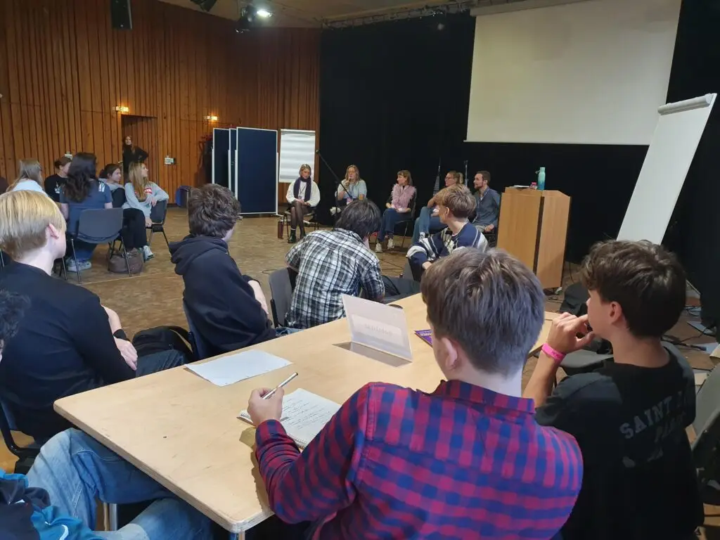 Schüler*innen sitzen an Tischen vor der Gesprächsrunde mit Politiker*innen.